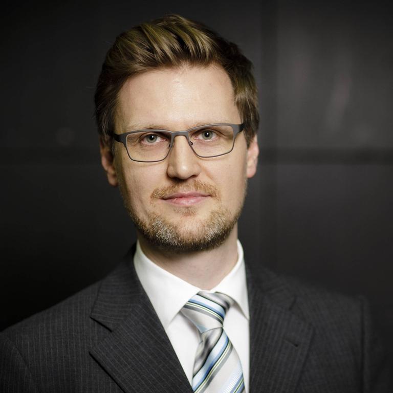 Martin Rücker trägt das Haar nach hinten gefönt und Dreitagebart. Er hat einen dunklen Anzug an, ein helles Hemd und eine helle Krawatte.