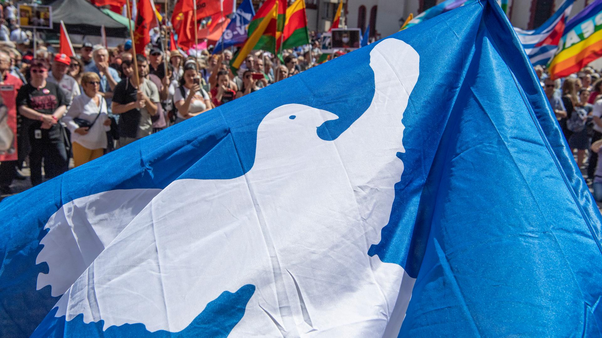 Auf einem Ostermarsch mit zahlreichen Teilnehmern ist eine blaue Fahne mit einer weißenn Friedenstaube zu sehen. Auf einem Ostermarsch mit zahlreichen Teilnehmern ist eine blaue Fahne mit einer weißenn Friedenstaube zu sehen.