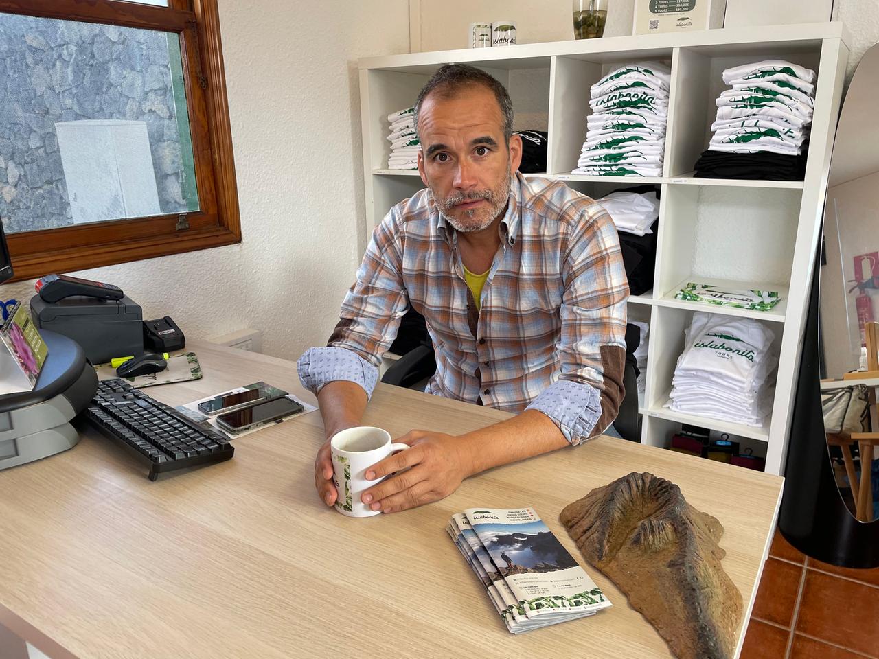 Ein Mann mit Dreitagebart sitzt an einem Tisch mit Computer vor einem Regal, die Kaffeetasse in der Hand. Ein Mann mit Dreitagebart sitzt an einem Tisch mit Computer vor einem Regal, die Kaffeetasse in der Hand.