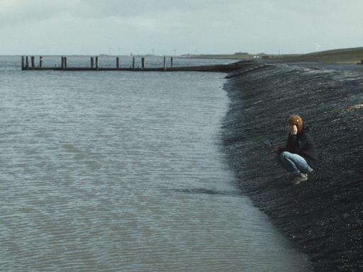 Ein Mädchen sitzt allein am Ufer des Meeres und schaut vor sich hin.