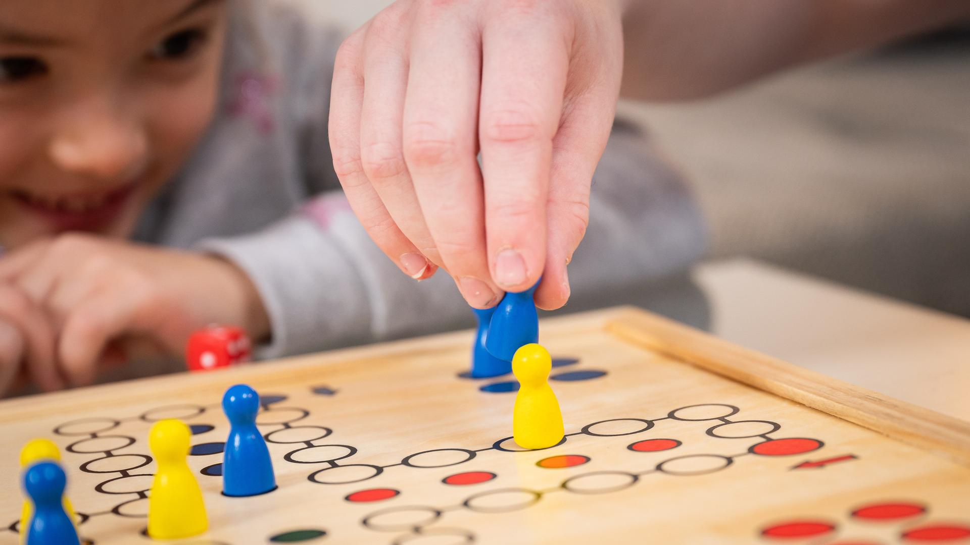 Nahaufnahme: Eine Kinderhand hält eine Spielfigur und schlägt damit eine andere aus dem Spiel "Mensch ärgere dich nicht" heraus, im Hintergrund sieht ein Mädchen gespannt zu und lacht. Nahaufnahme: Eine Kinderhand hält eine Spielfigur und schlägt damit eine andere aus dem Spiel "Mensch ärgere dich nicht" heraus, im Hintergrund sieht ein Mädchen gespannt zu und lacht.