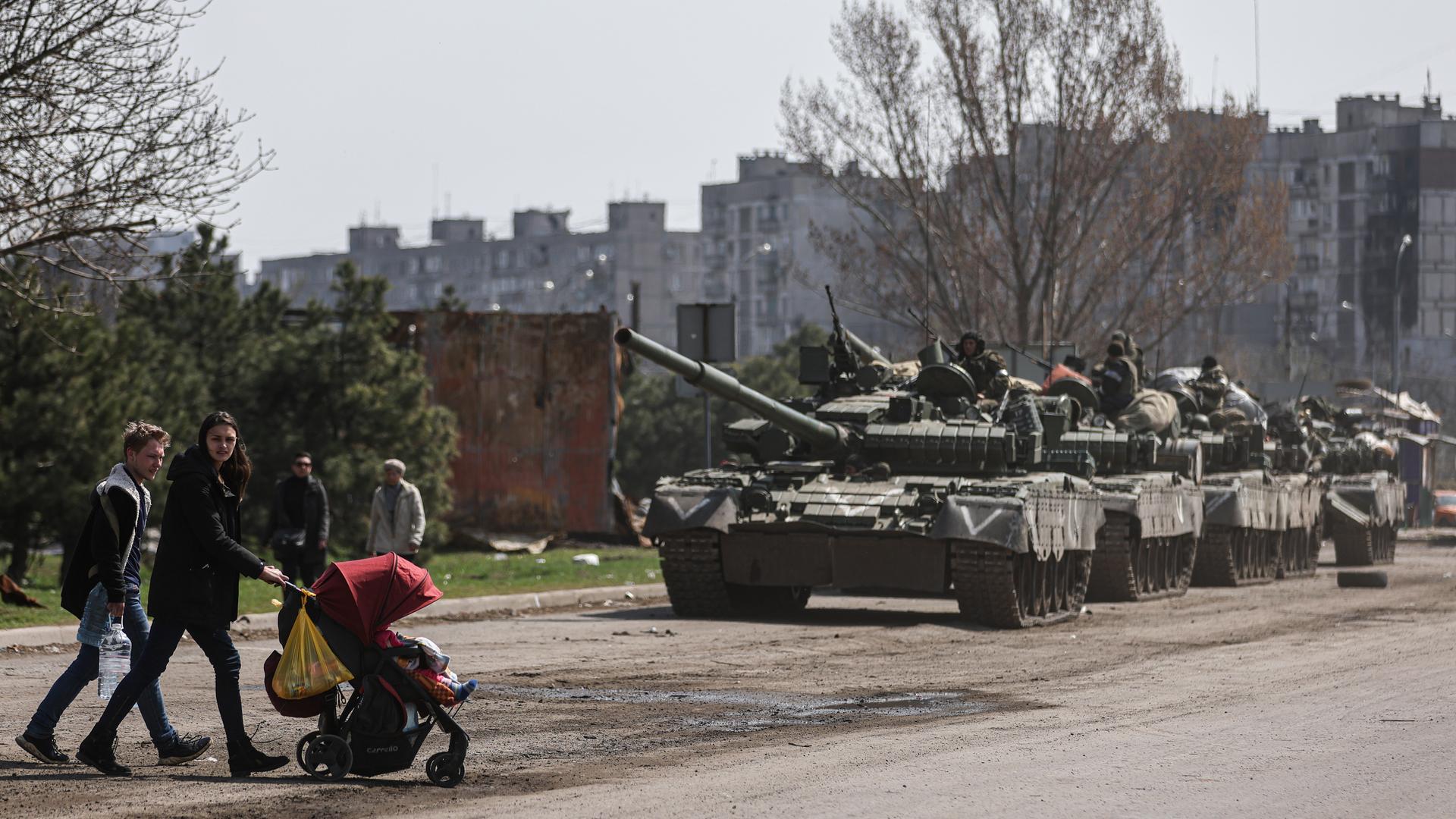 Mariupol im April 2022: Zivilisten mit Kinderwagen überqueren die Straße vor mehreren russischen Panzern Mariupol im April 2022: Zivilisten mit Kinderwagen überqueren die Straße vor mehreren russischen Panzern