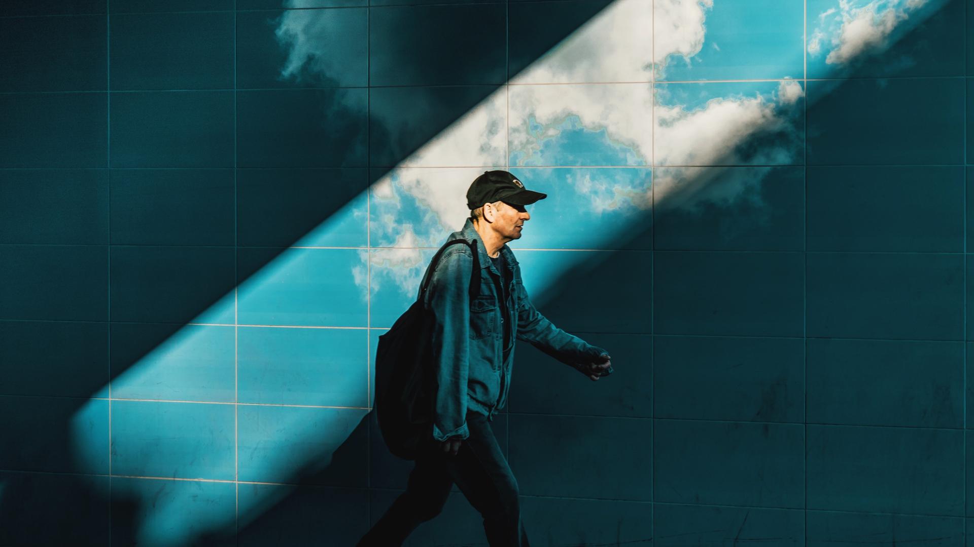 Ein Mann vor blauen Fliesen, wie in einem U-Bahn-Schacht. Gehend, eine Kappe auf dem Kopf. Ein breiter Lichtstrahl zieht sich von rechts oben nach links unten durch das Bild und erhellt den Mann und die Fliesen. Darauf sind Wolken zu sehen. Ein Mann vor blauen Fliesen, wie in einem U-Bahn-Schacht. Gehend, eine Kappe auf dem Kopf. Ein breiter Lichtstrahl zieht sich von rechts oben nach links unten durch das Bild und erhellt den Mann und die Fliesen. Darauf sind Wolken zu sehen.