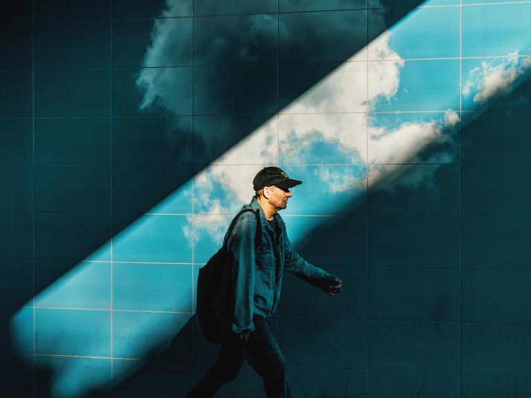 Ein Mann vor blauen Fliesen, wie in einem U-Bahn-Schacht. Gehend, eine Kappe auf dem Kopf. Ein breiter Lichtstrahl zieht sich von rechts oben nach links unten durch das Bild und erhellt den Mann und die Fliesen. Darauf sind Wolken zu sehen.