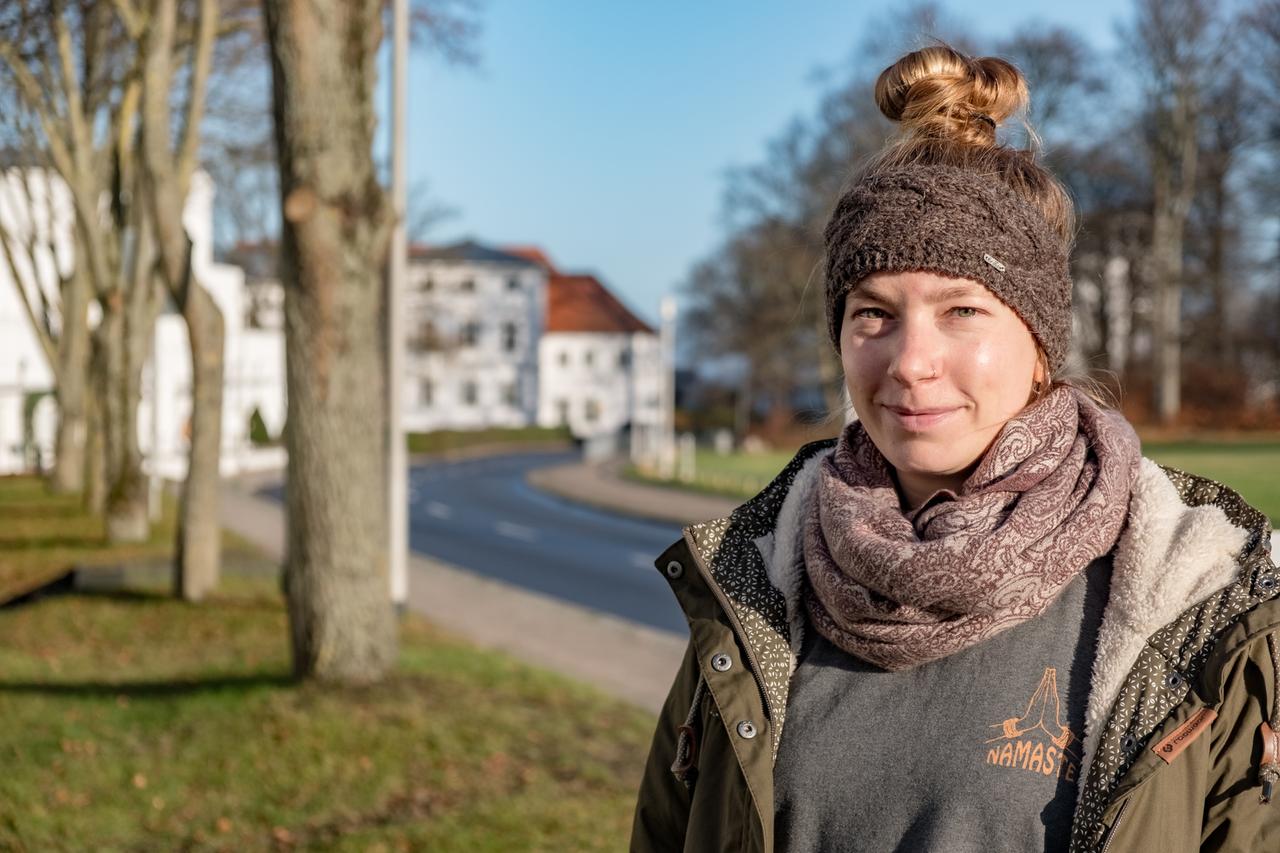 Katharina Sozialarbeiterin aus Hannover steht vor einer Bushaltestelle mit Handgepäck.