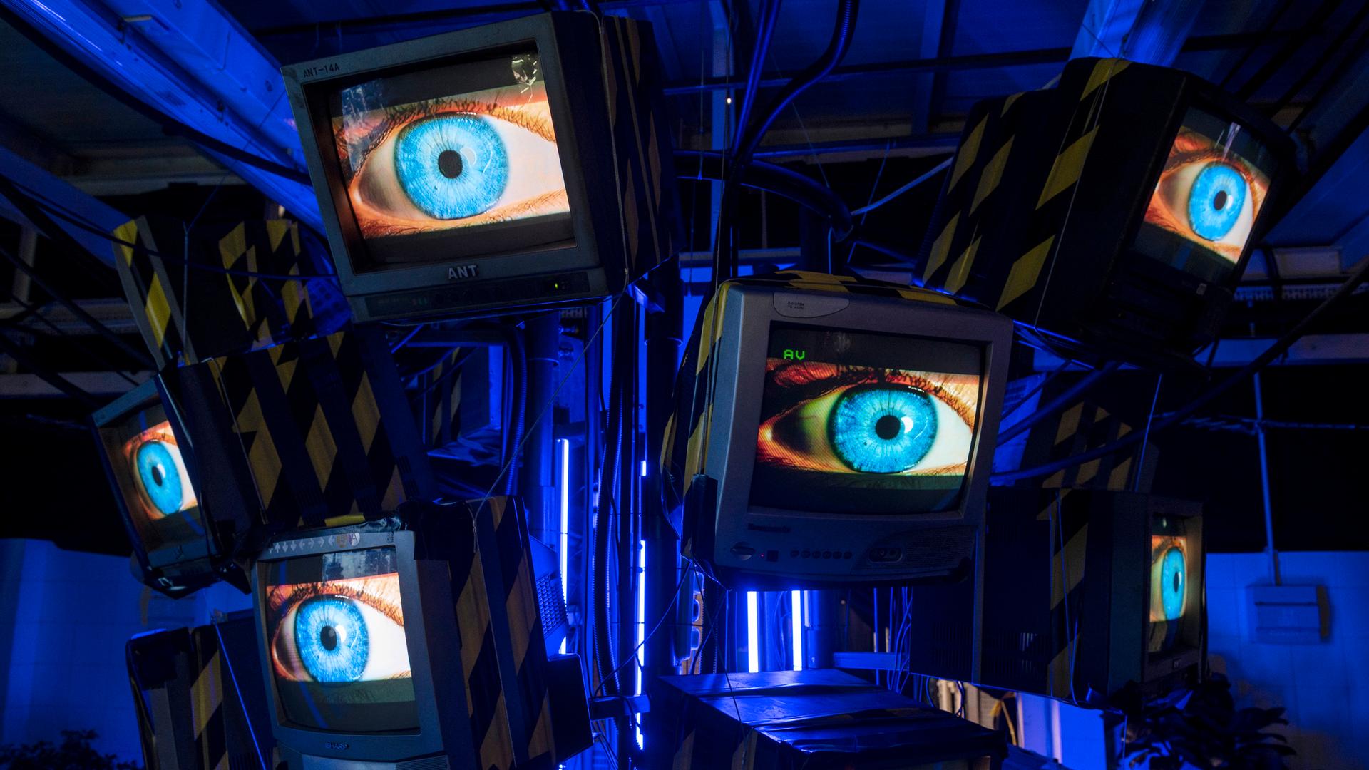 Eine Installation von acht Fernsehbildschirmen, die in einem Halbkreis angeordnet sind, zeigen jeweils die gleiche Nahaufnahme eines menschlichen Auges mit einer blauen Iris.
