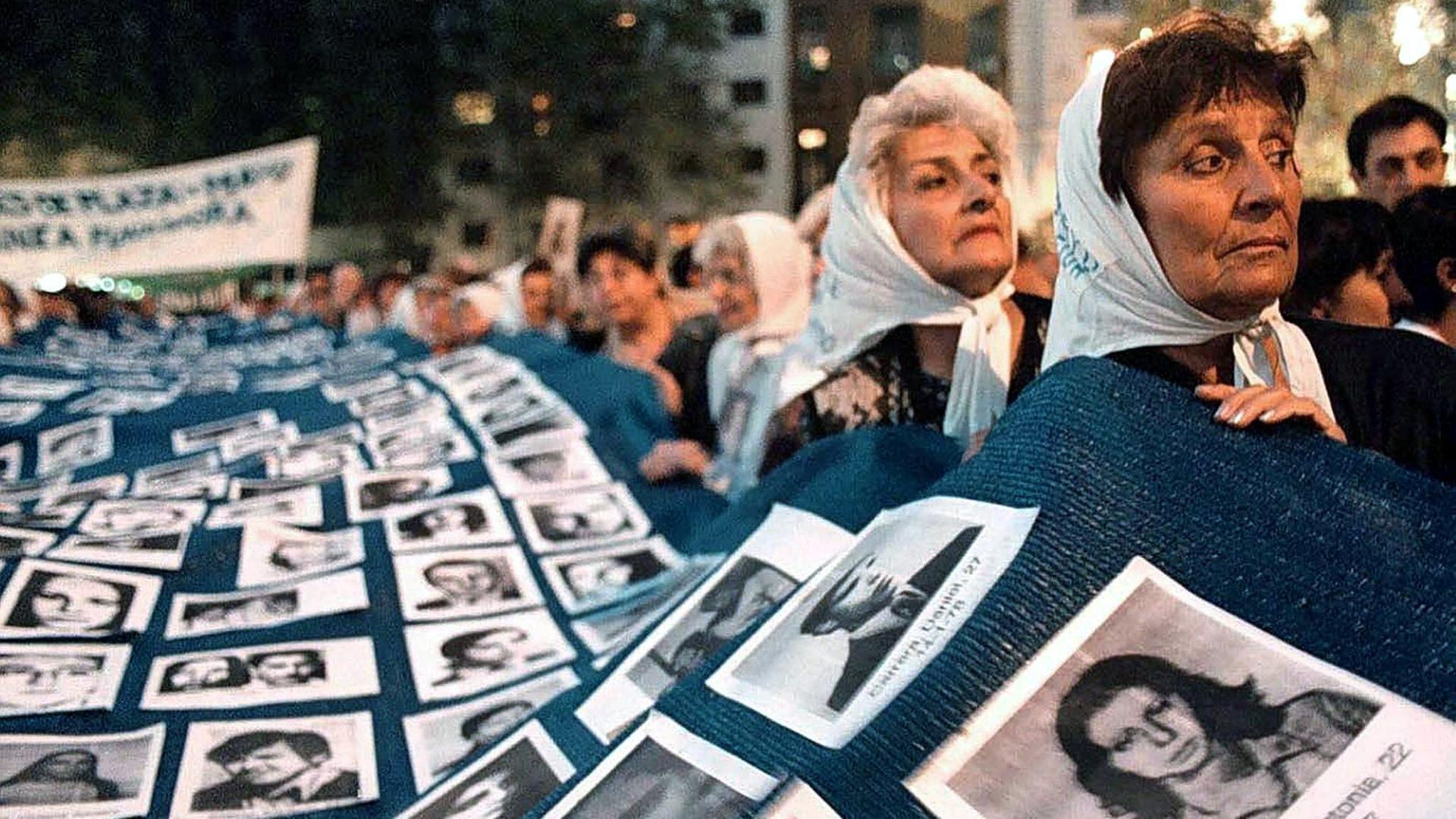 Frauen tragen eine große Decke, auf der Fotos von vermissten Menschen befestigt sind. 