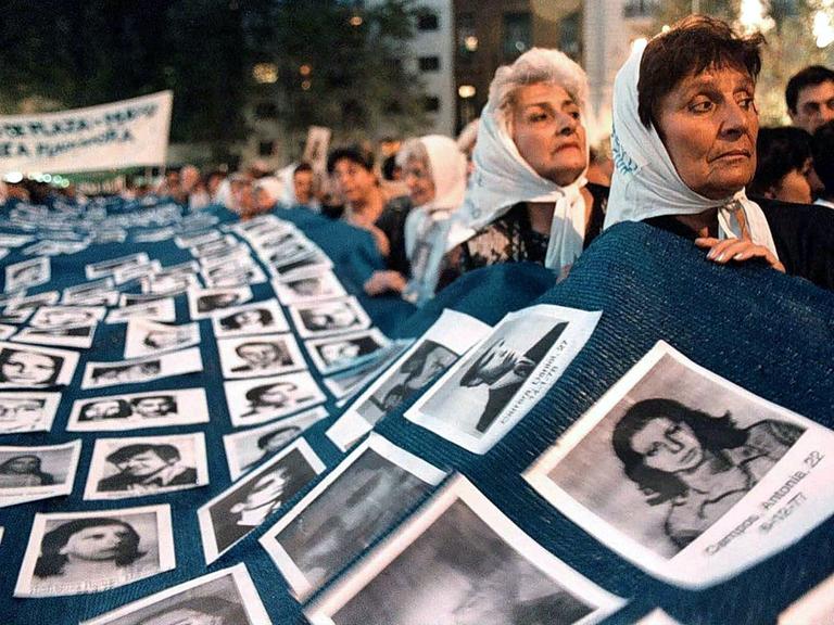 Frauen tragen eine große Decke, auf der Fotos von vermissten Menschen befestigt sind. 