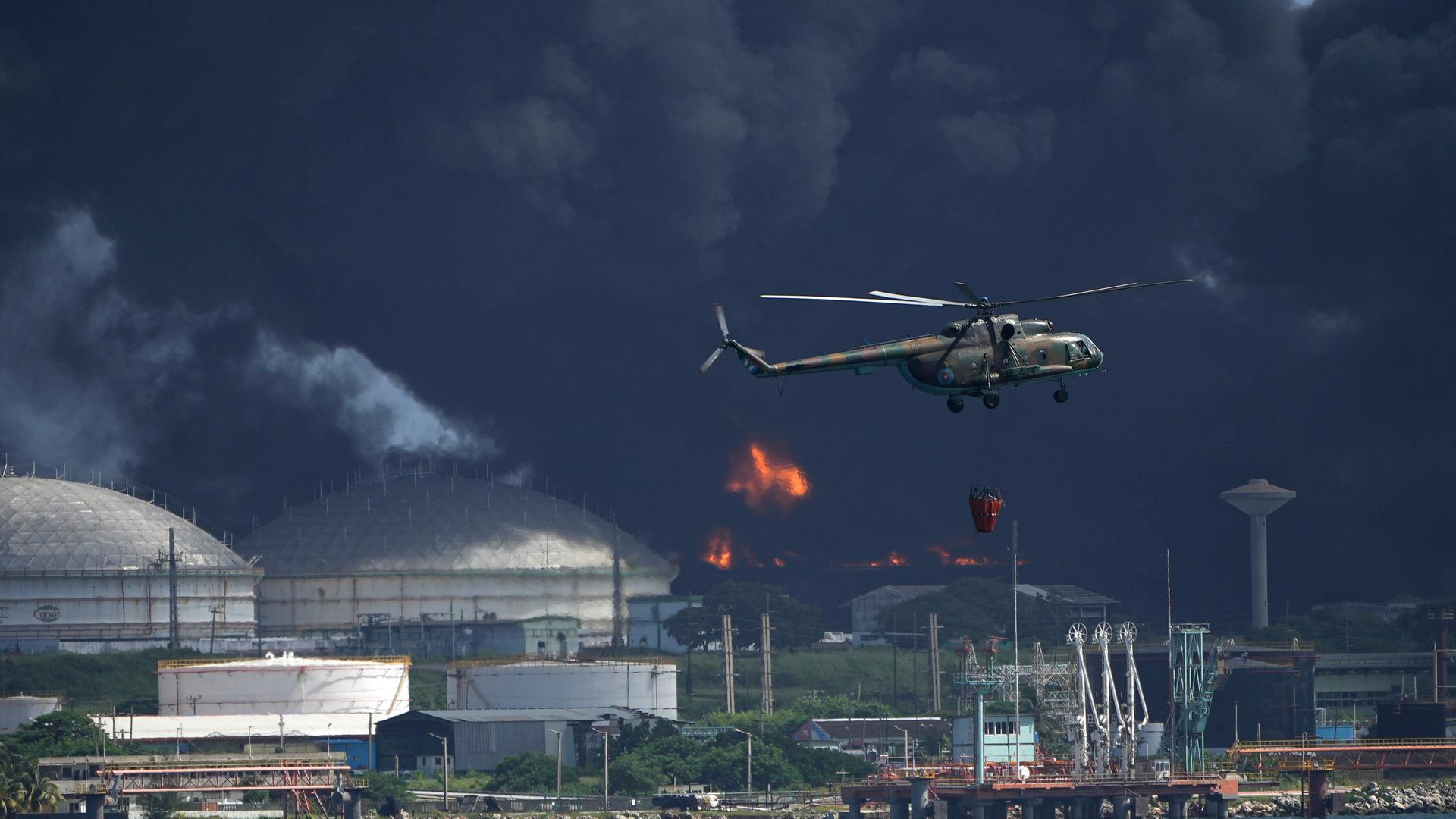 Ein mit Wasser beladener Hubschrauber fliegt über die Supertanker-Basis Matanzas, von der dunkler Rauch und Flammen aufsteigen, während Feuerwehrleute versuchen, den Brand zu löschen.