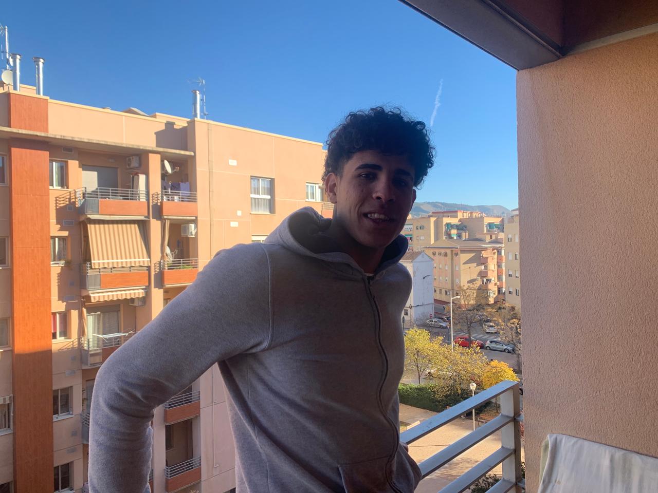 Ein junger Mann im grauen Sweater steht vor dem Hintergrund eines blauen Himmels auf dem Balkon eines Häuserblocks Ein junger Mann im grauen Sweater steht vor dem Hintergrund eines blauen Himmels auf dem Balkon eines Häuserblocks