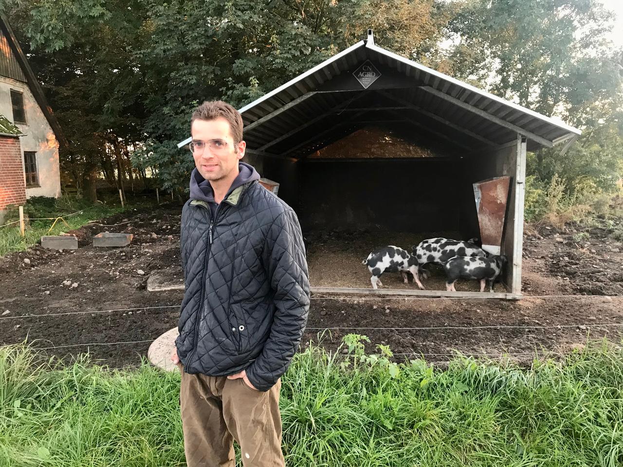 Ein Mann in dunklem Anorak steht vor einem Stall mit scheckigen Schweinen. Ein Mann in dunklem Anorak steht vor einem Stall mit scheckigen Schweinen.
