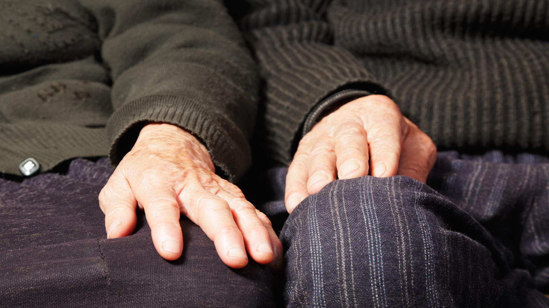 Ein älteres Paar sitzt auf dem Sofa. Nahaufnahme der Hände, die nebeneinander jeweils auf den Knien ruhen.