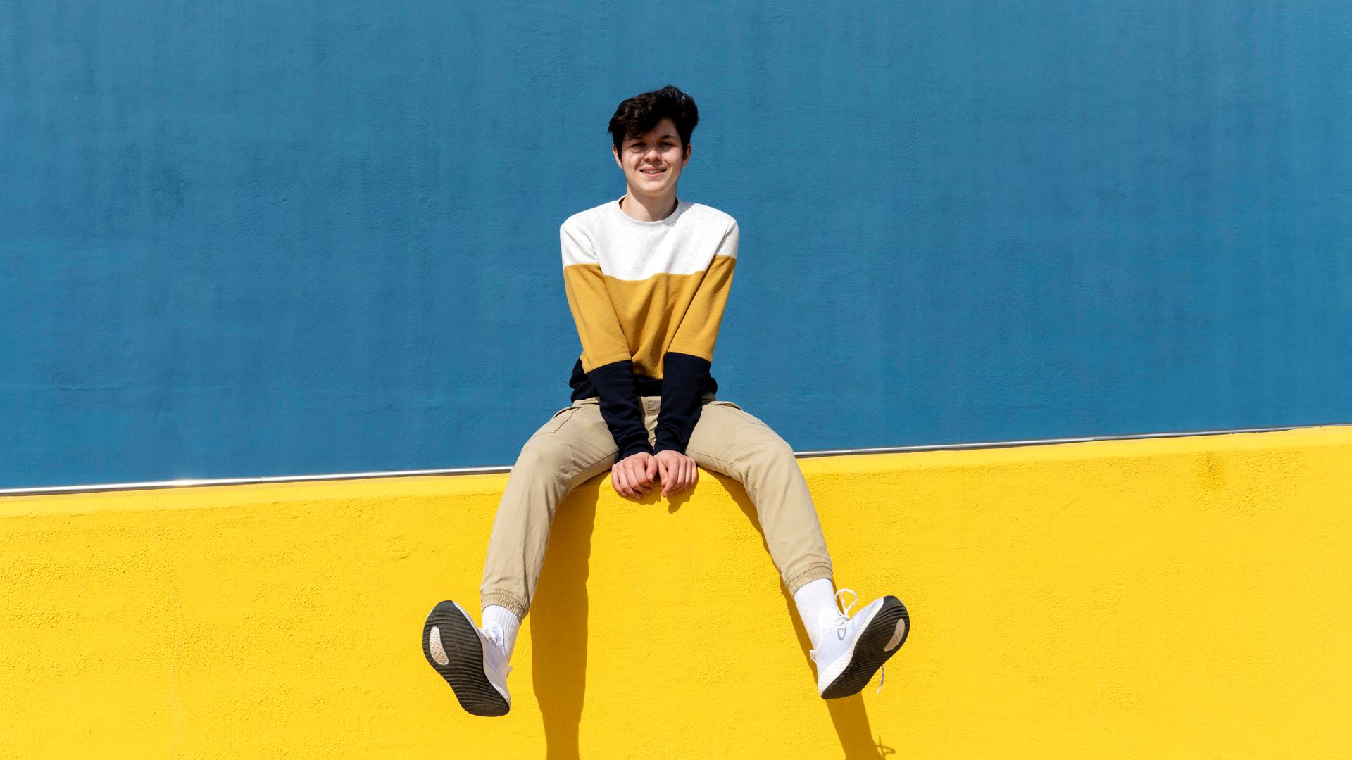 Ein zuversichtlich schauender junger Mann sitzt auf einer gelben Mauer.