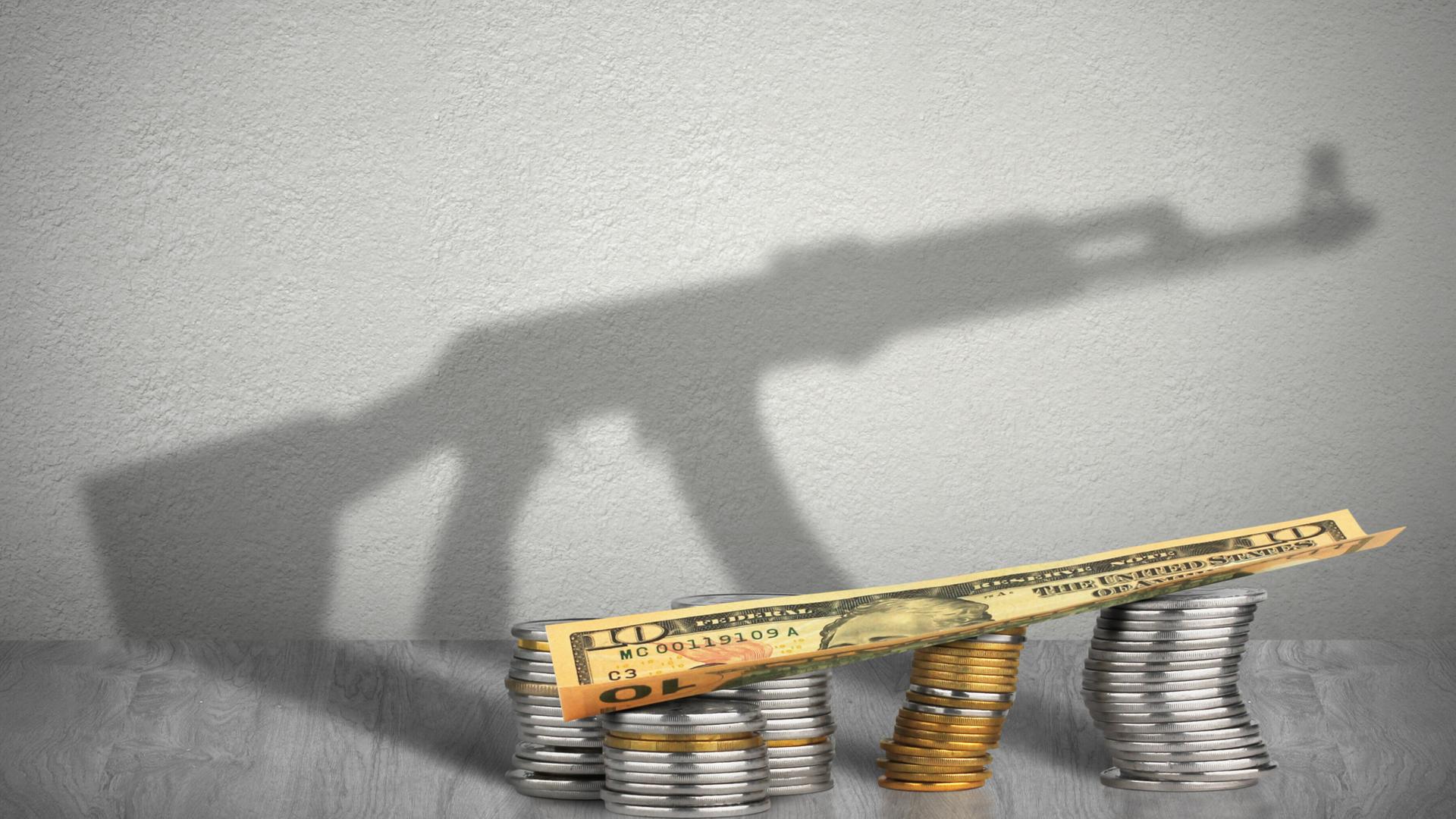 Im Vordergrund sind auf dem Symbolbild Münztürme zu sehen, auf denen eine Banknote liegt. Im Hintergrund ist ein Schatten eines Maschinengewehrs zu sehen. Auf den ersten Blick scheint der MG-Schatten der Schattev der Münztürme und der Banknote zu sein. Im Vordergrund sind auf dem Symbolbild Münztürme zu sehen, auf denen eine Banknote liegt. Im Hintergrund ist ein Schatten eines Maschinengewehrs zu sehen. Auf den ersten Blick scheint der MG-Schatten der Schattev der Münztürme und der Banknote zu sein.