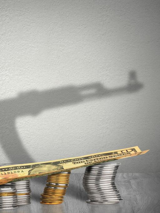 Im Vordergrund sind auf dem Symbolbild Münztürme zu sehen, auf denen eine Banknote liegt. Im Hintergrund ist ein Schatten eines Maschinengewehrs zu sehen. Auf den ersten Blick scheint der MG-Schatten der Schattev der Münztürme und der Banknote zu sein.