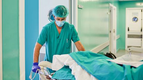 Ein Mann in grünem Schutzanzug, Haube und Maske beugt sich über einen Patienten, der in einem Krankenhausbett liegt.