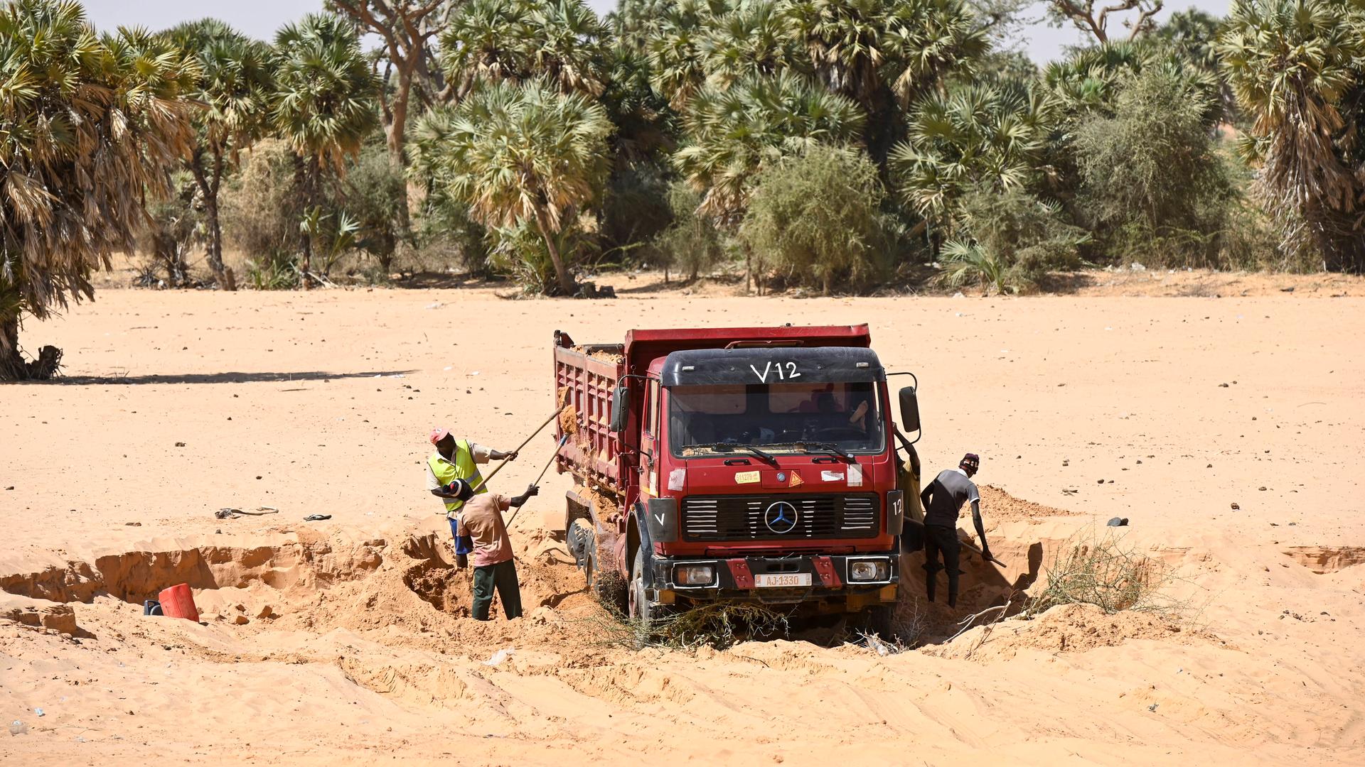 Ein Lastwagen auf sandigem Untergrund, Arbeiter schaufeln Sand in das Fahrzeug. Im Hintergrund sind Bäume zu sehen. Ein Lastwagen auf sandigem Untergrund, Arbeiter schaufeln Sand in das Fahrzeug. Im Hintergrund sind Bäume zu sehen.