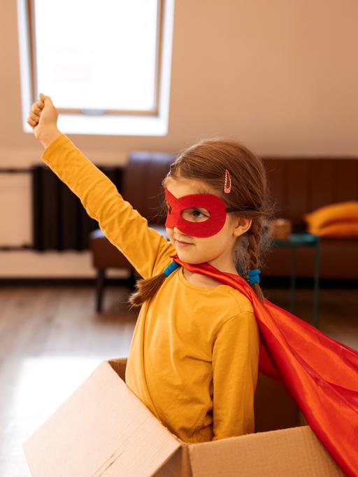 Ein Kind in einem Pappkarton und Heldinnenkostum reckt die Faust in die Höhe.