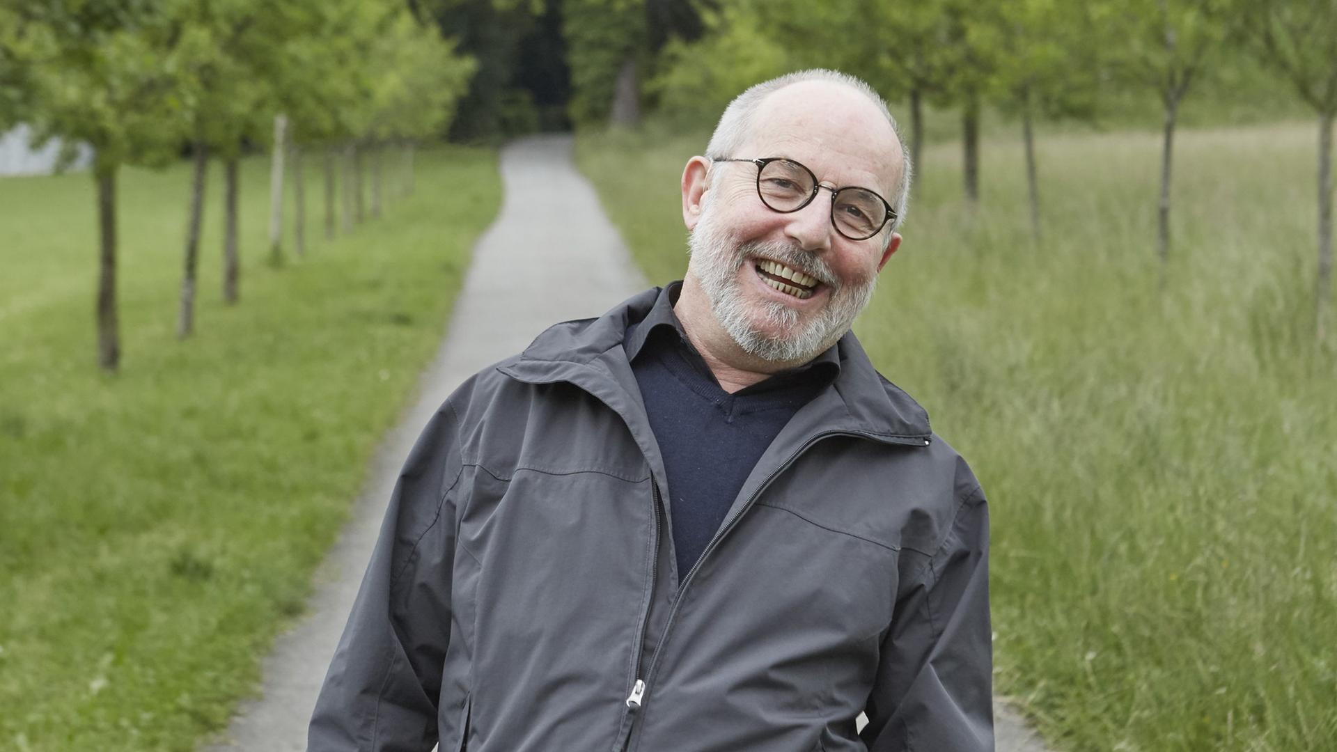 Der Schweizer Schriftsteller Thomas Hürlimann steht auf einem von Bäumen gesäumten schmalen Weg und lacht. Rechts und links sind grüne Wiesen zu sehen. Hürlimann trägt eine graue Halbglatze und Brille.