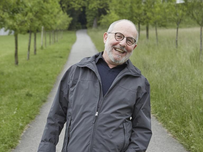 Der Schweizer Schriftsteller Thomas Hürlimann steht auf einem von Bäumen gesäumten schmalen Weg und lacht. Rechts und links sind grüne Wiesen zu sehen. Hürlimann trägt eine graue Halbglatze und Brille.