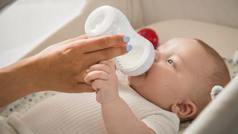 Eine Hand hält die Flasche, aus dem eine in weiß gekleidetes Baby trinkt. Es liegt in einem Babybett.