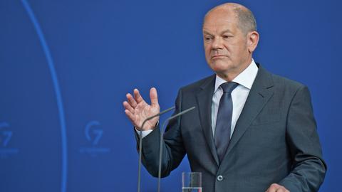 Bundeskanzler Olaf Scholz hebt seine rechte Hand und steht am Pult vor zwei Mikrofonen während einer Pressekonferenz.