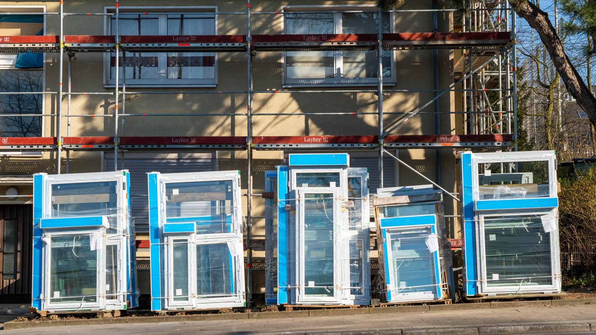 Vor einem eingerüsteten älteren Mehrfamilienhaus stehen neue Fenster und Bauelemente für eine Wärmedämmung aufgereiht. Vor einem eingerüsteten älteren Mehrfamilienhaus stehen neue Fenster und Bauelemente für eine Wärmedämmung aufgereiht.