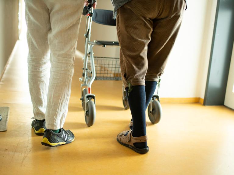 Eine Person begleitet einen Mann mit Rollator im Hausflur. Es sind vor allem die Beine und Füsse zu sehen.