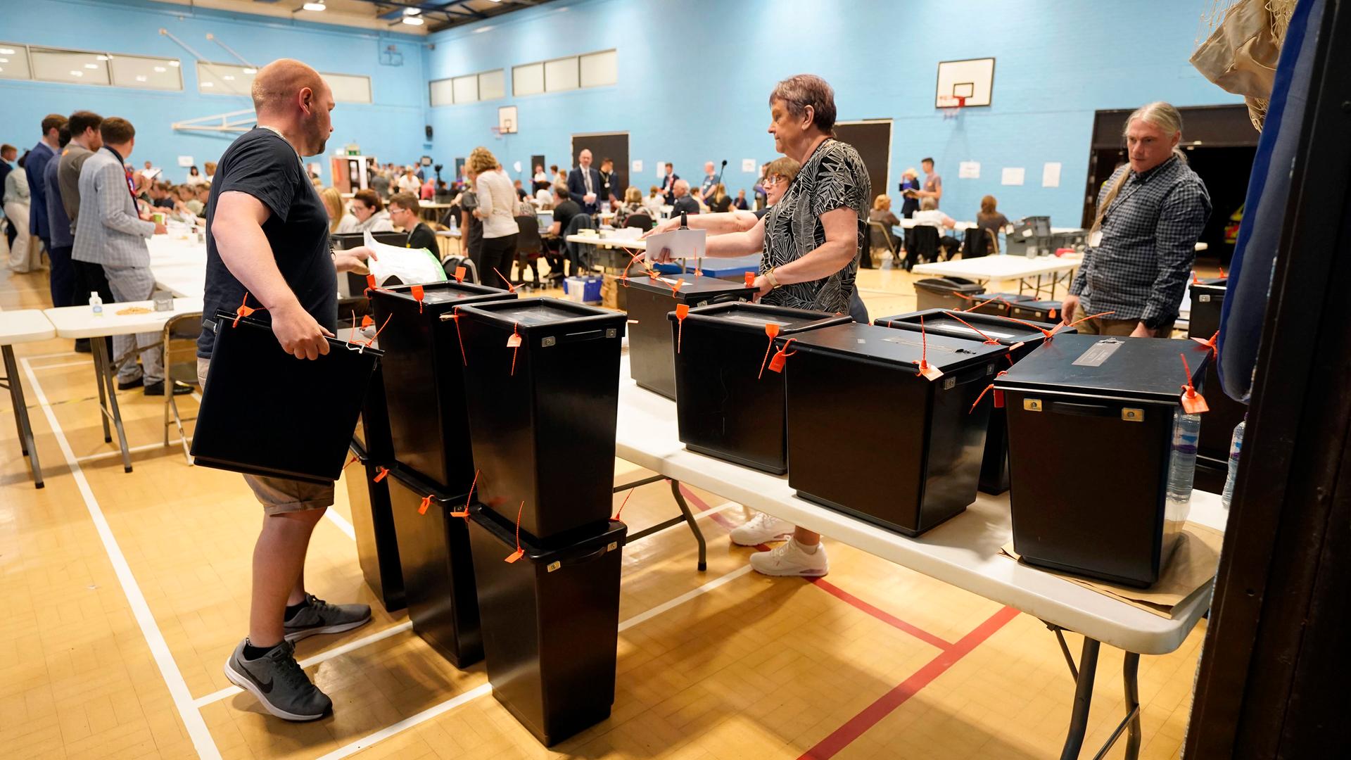 Menschen stehen in einer Turnhalle vor schwarzen Wahlurnen. Menschen stehen in einer Turnhalle vor schwarzen Wahlurnen.