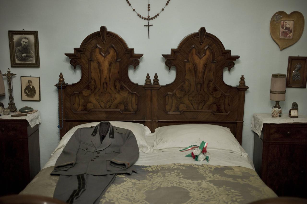 Ein Doppelbett. Auf der einen Seite liegt ein gebügelter Anzug mit Abzeichen. An den Wänden Fotos und ein Kreuz. Ein Doppelbett. Auf der einen Seite liegt ein gebügelter Anzug mit Abzeichen. An den Wänden Fotos und ein Kreuz.