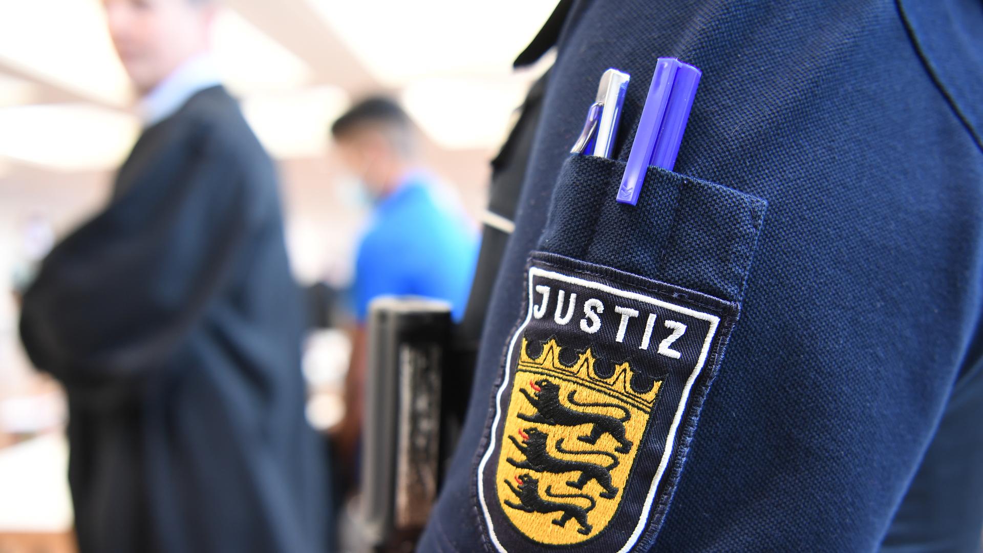 Ein Angeklagter steht im Juli 2020 im Gerichtssaal in der Messe Ulm, während im Vordergrund eine Justizmitarbeiterin steht, deren Justiz-Logo am Ärmel zu sehen ist. Fünf Männer sind im Juli 2020 wegen Vergewaltigung einer 14-Jährigen angeklagt. Ein Angeklagter steht im Juli 2020 im Gerichtssaal in der Messe Ulm, während im Vordergrund eine Justizmitarbeiterin steht, deren Justiz-Logo am Ärmel zu sehen ist. Fünf Männer sind im Juli 2020 wegen Vergewaltigung einer 14-Jährigen angeklagt.