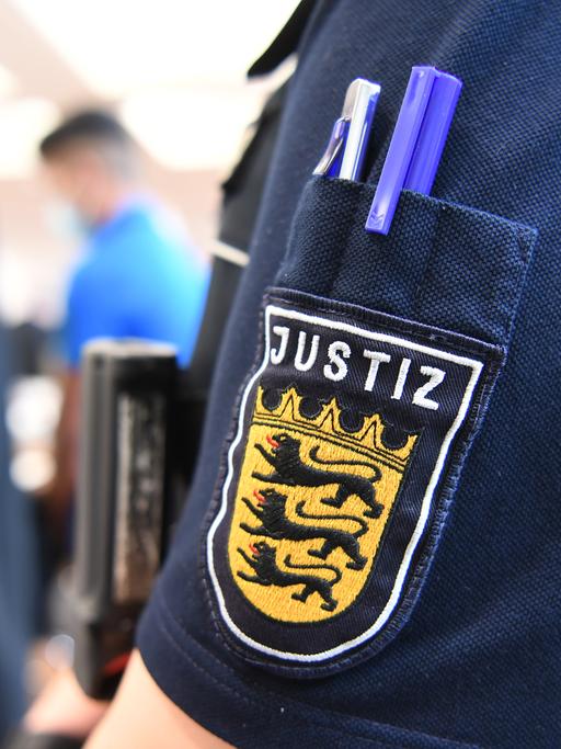 Ein Angeklagter steht im Juli 2020 im Gerichtssaal in der Messe Ulm, während im Vordergrund eine Justizmitarbeiterin steht, deren Justiz-Logo am Ärmel zu sehen ist. Fünf Männer sind im Juli 2020 wegen Vergewaltigung einer 14-Jährigen angeklagt.