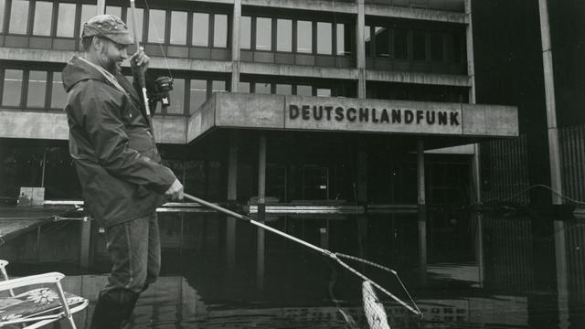 Schwarz-Weiß-Aufnahme eines Mannes, der vor dem Funkhaus des Deutschlandfunk in Köln eine Angel ins Wasser hält Schwarz-Weiß-Aufnahme eines Mannes, der vor dem Funkhaus des Deutschlandfunk in Köln eine Angel ins Wasser hält