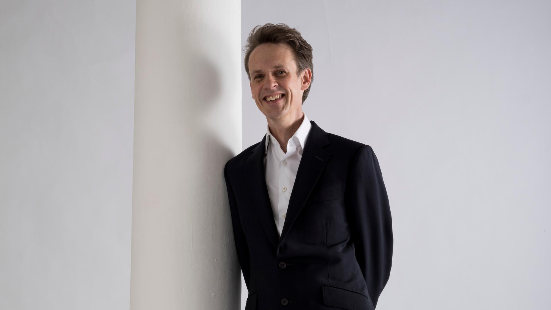 Ein Mann lehnt gegen eine weiße Säule. Er trägt einen schwarzen Anzug und lächelt. Es ist der Tenor Ian Bostridge. Ein Mann lehnt gegen eine weiße Säule. Er trägt einen schwarzen Anzug und lächelt. Es ist der Tenor Ian Bostridge.