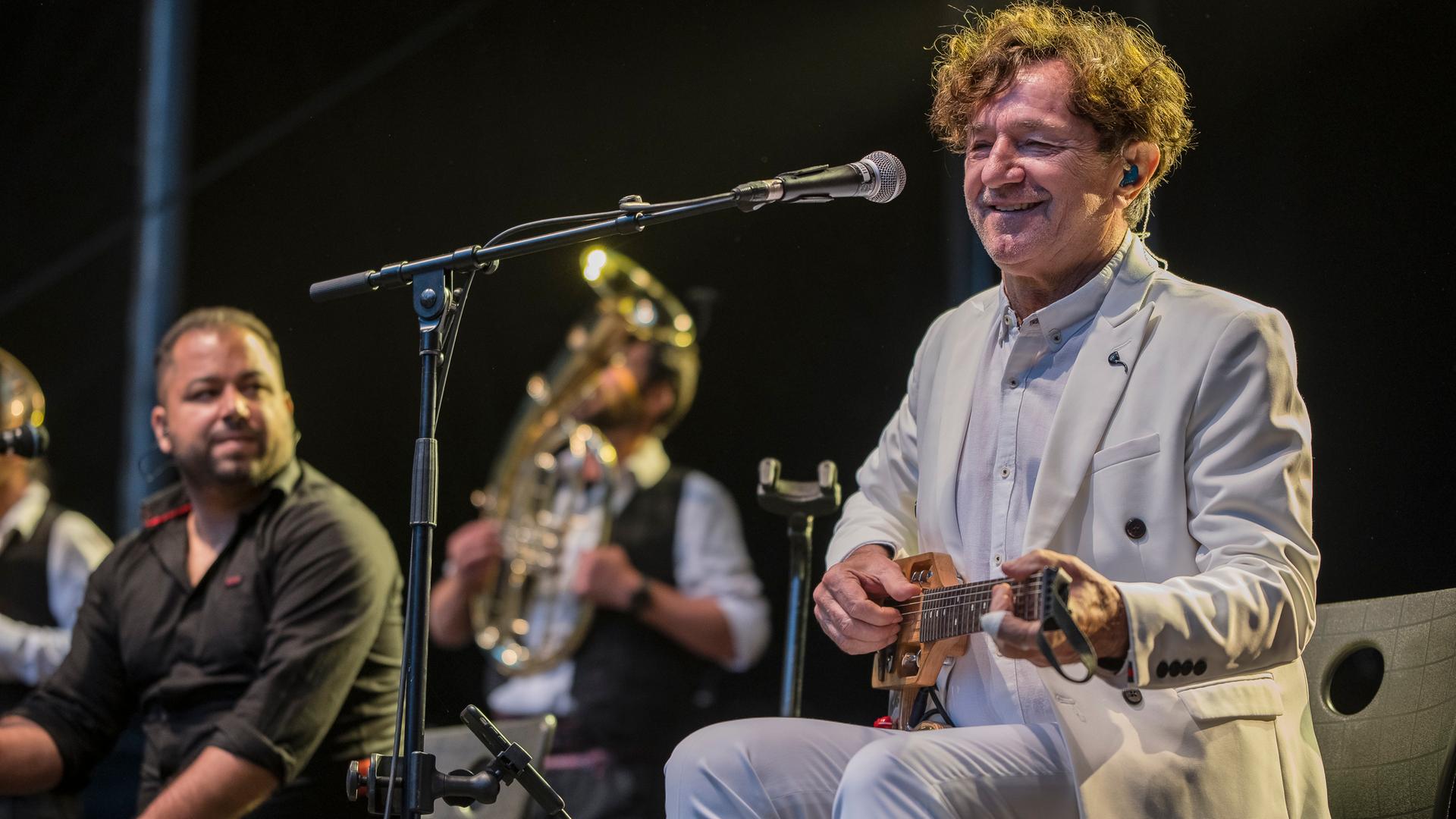 Ein Mann in einem hellen Anzug sitzt auf einem Stuhl, auf dem Schoß hält er eine kleine Gitarre. Vor ihm steht ein Mikrofon. Er hat kurze braune, gelockte Haare und blickt freundlich. Ein Mann in einem hellen Anzug sitzt auf einem Stuhl, auf dem Schoß hält er eine kleine Gitarre. Vor ihm steht ein Mikrofon. Er hat kurze braune, gelockte Haare und blickt freundlich.