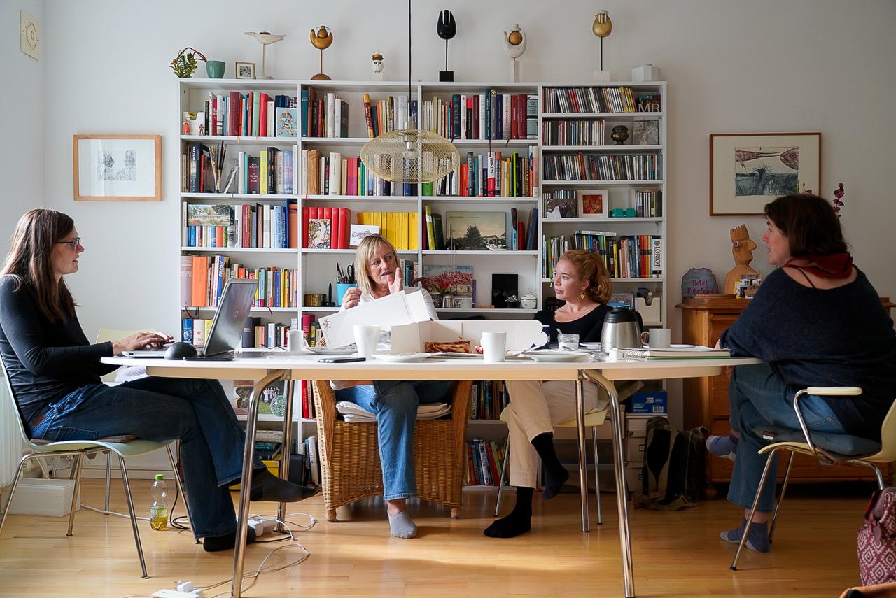 Vier Frauen sitzen in einem Büro mit Bücherregalen an einem langen Konferenztisch. Vier Frauen sitzen in einem Büro mit Bücherregalen an einem langen Konferenztisch.