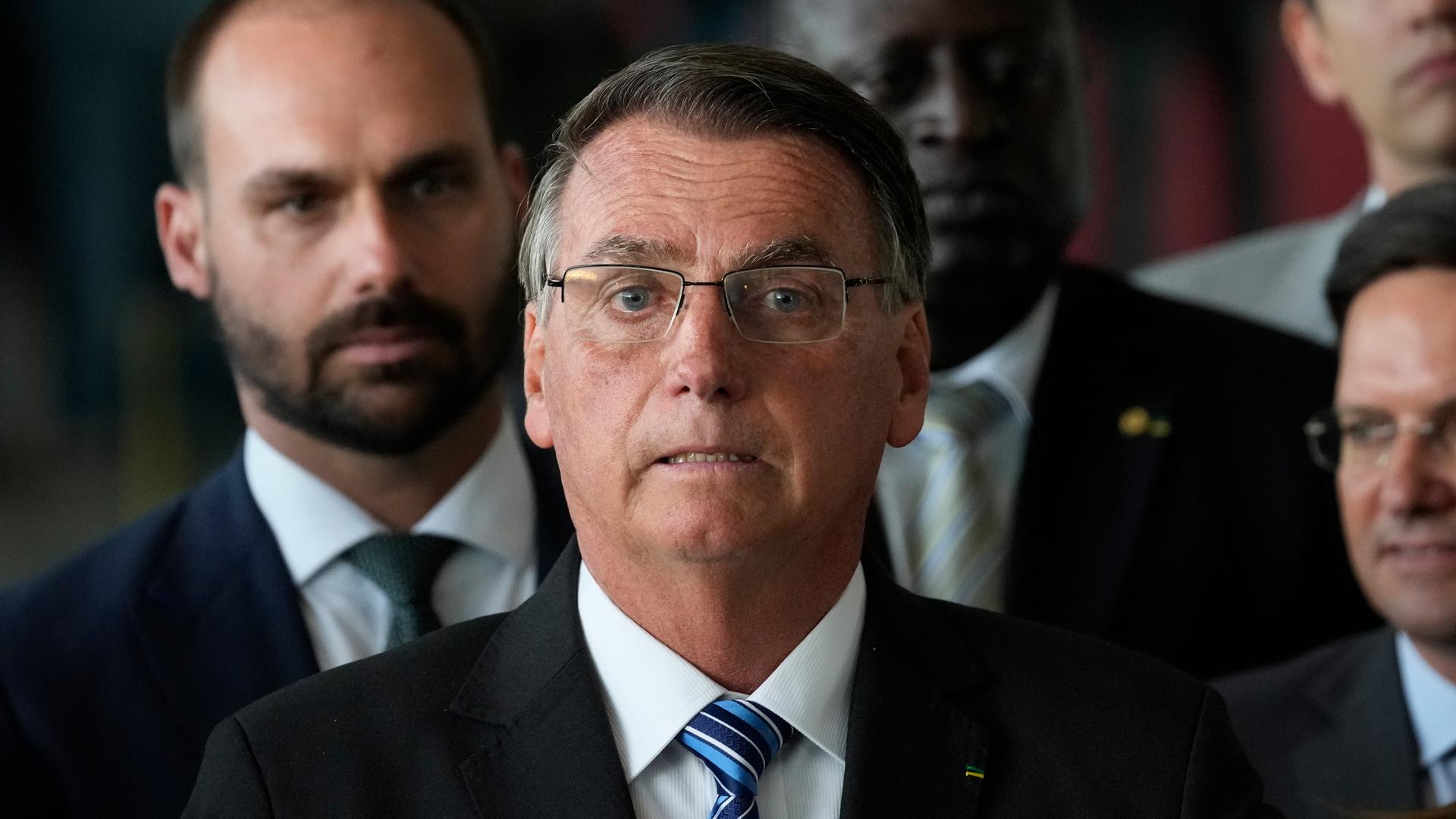 Großaufnahme von Jair Bolsonaro, der betreten mit halb geöffnetem Mund in die Kamera schaut. Im Hintergrund sind Mitglieder seiner Regierung zu sehen. Großaufnahme von Jair Bolsonaro, der betreten mit halb geöffnetem Mund in die Kamera schaut. Im Hintergrund sind Mitglieder seiner Regierung zu sehen.