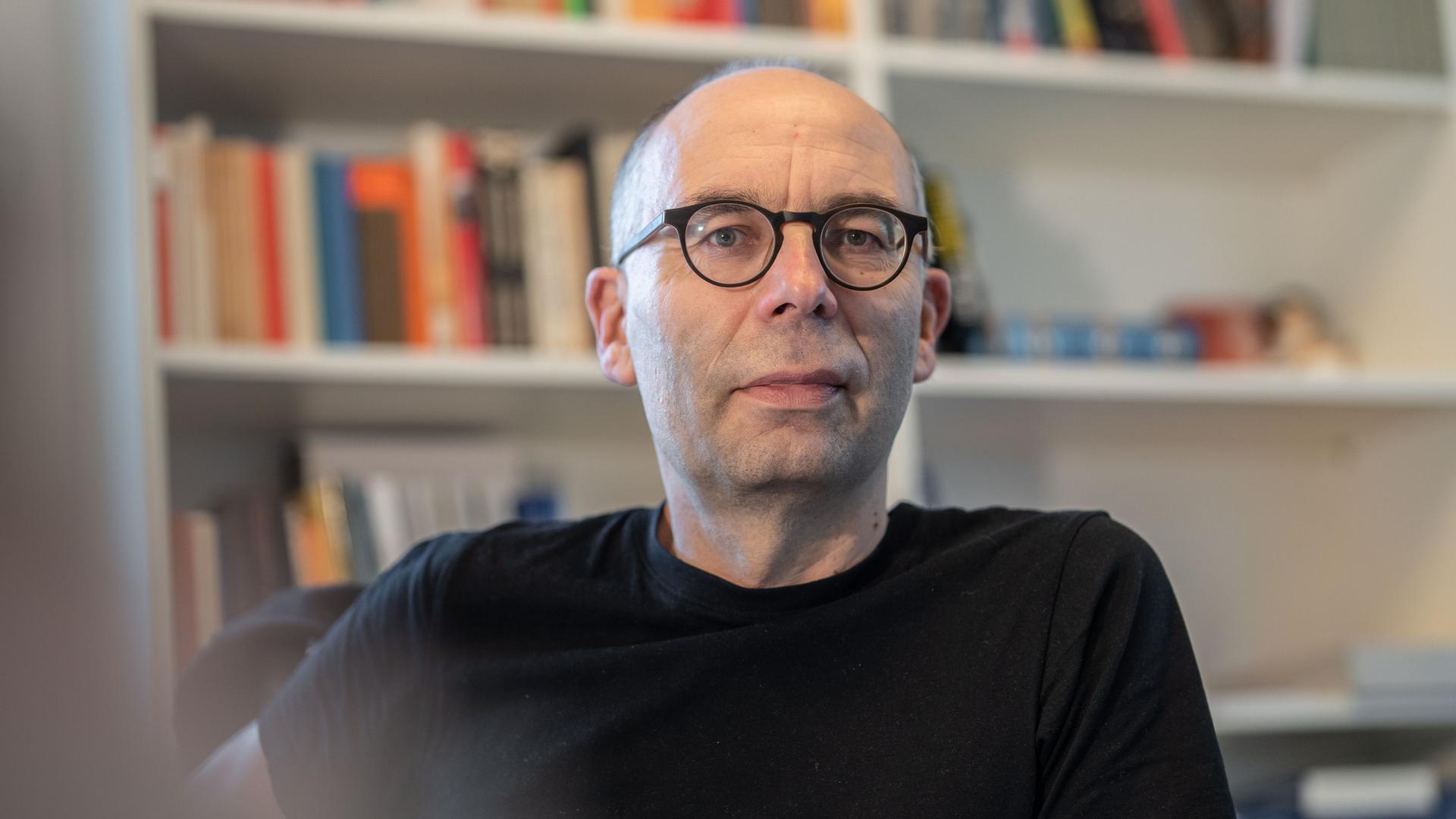 Der Soziologe Stephan Lessenich sitzt in einem schwarzen T-Shirt an seinem Schreibtisch vor einem Bücherregal.