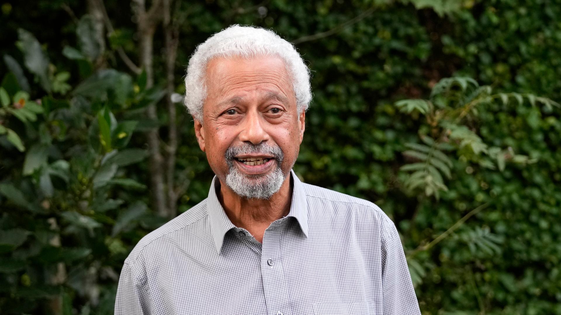 Der Literaturnobelpreisträger Abdulrazak Gurnah steht vor einem Pflanzendickicht mit den Händen in den Hosentaschen und lächelt in die Kamera. Der Literaturnobelpreisträger Abdulrazak Gurnah steht vor einem Pflanzendickicht mit den Händen in den Hosentaschen und lächelt in die Kamera.