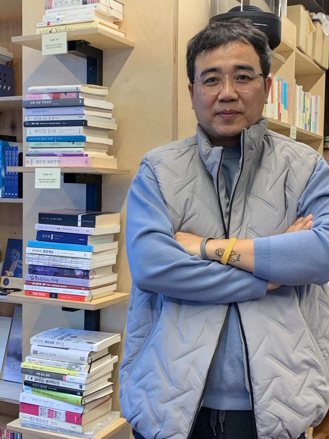 Choi Joo Kwang steht mit verschränkten Armen neben einem Bücherregal in seinem Café. Choi Joo Kwang steht mit verschränkten Armen neben einem Bücherregal in seinem Café.