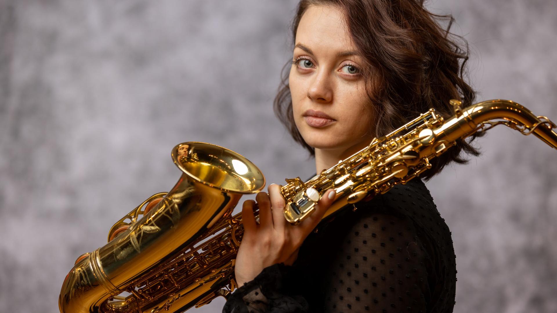 Eine junge Frau mit braunen Haaren dreht ihren Kopf seitwärts und blickt in die Kamera. In ihrer linken Hand hält sie ein Saxofon, das Instrument lehnt auf ihrer linken Schulter. Eine junge Frau mit braunen Haaren dreht ihren Kopf seitwärts und blickt in die Kamera. In ihrer linken Hand hält sie ein Saxofon, das Instrument lehnt auf ihrer linken Schulter.