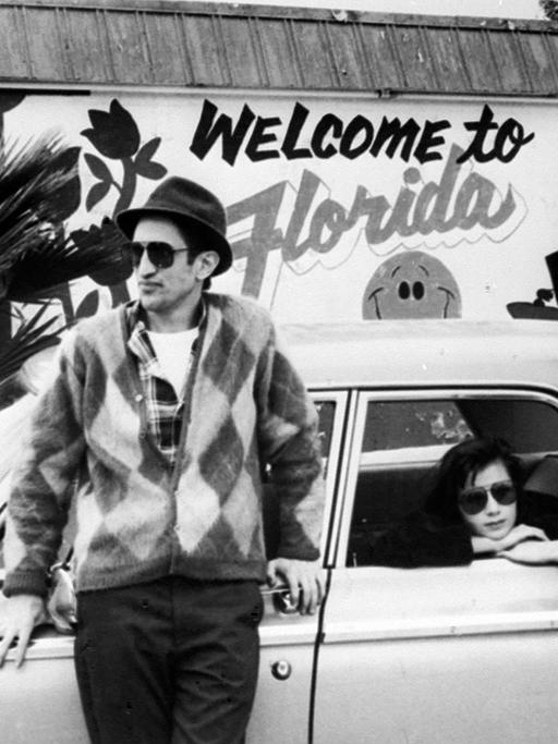 Zwei Männer lehnen an einem Auto, in dem eine Frau sitzt und aus dem Fenster schaut. Im Hintergrund steht auf einer Wand "Welcome to Florida"