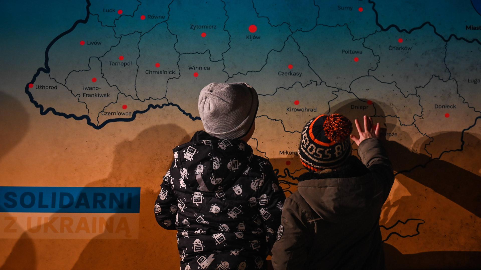Zwei Kinder sind von hinten vor einer Karte der Ukraine zu sehen, sie zeigen auf die Karte