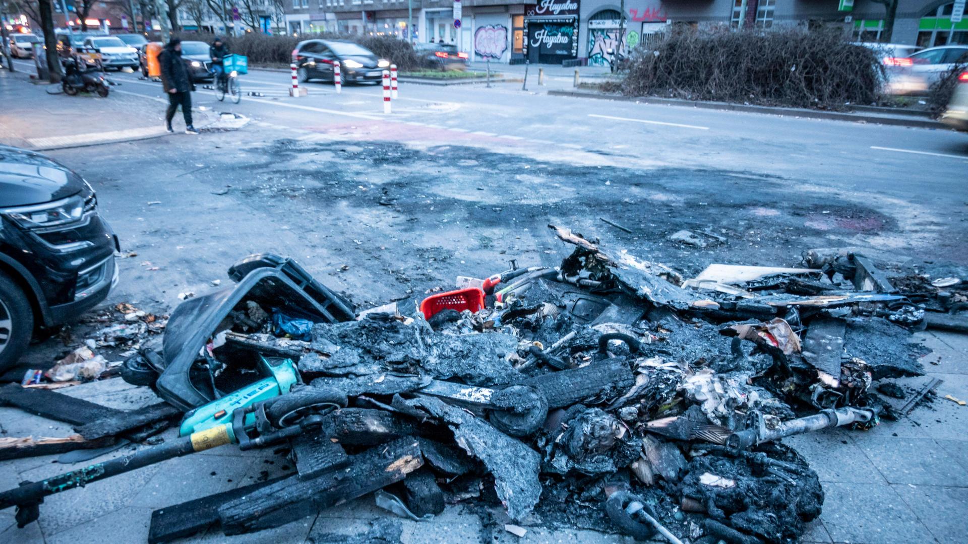 Überreste von verbrannten Mülltonnen und E-Scootern auf einer Straße.