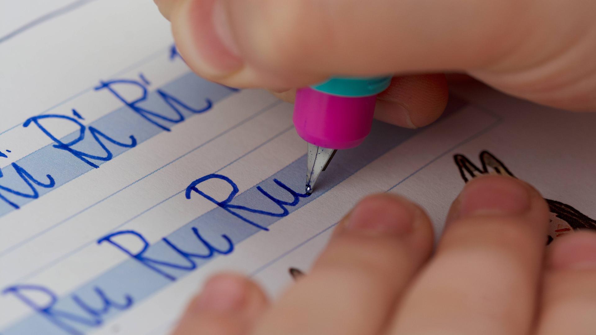 Eine Kinderhand mit Stift schreibt in ein Heft "Ri Ri Ri" und darunter "Ru Ru Ru".