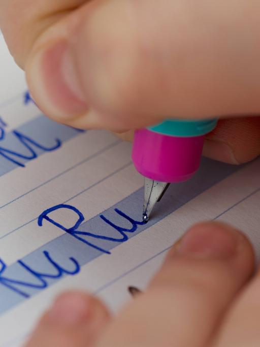 Eine Kinderhand mit Stift schreibt in ein Heft "Ri Ri Ri" und darunter "Ru Ru Ru".