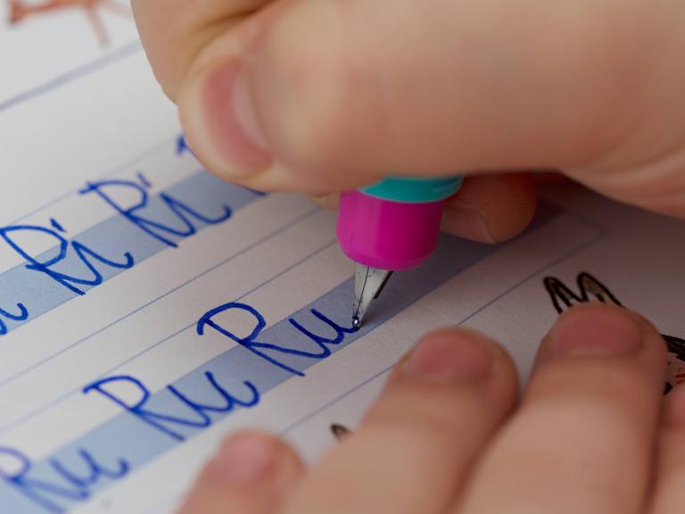 Eine Kinderhand mit Stift schreibt in ein Heft "Ri Ri Ri" und darunter "Ru Ru Ru".