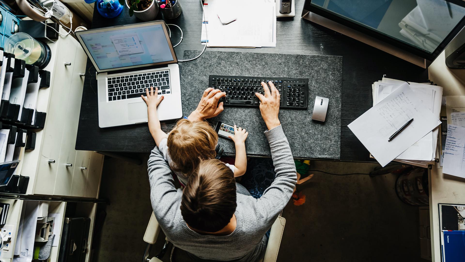 Eine Mutter sitzt mit einem Kind auf dem Schoß am Schreibtisch und versucht an einem Computer zu arbeiten. Eine Mutter sitzt mit einem Kind auf dem Schoß am Schreibtisch und versucht an einem Computer zu arbeiten.