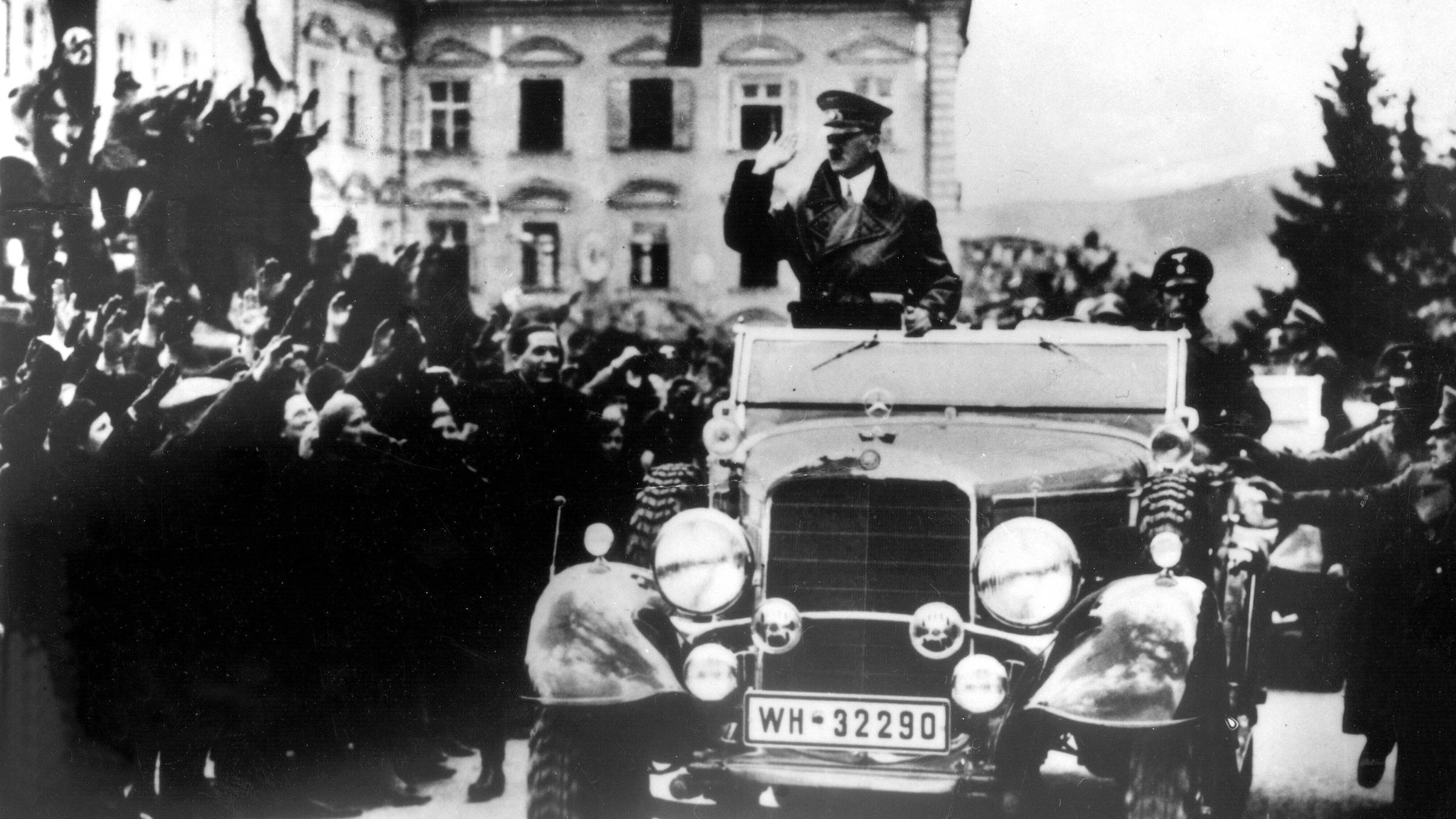 Historische schwarz-weiß-Aufnahme zeigt Adolf Hitler im offenen Fahrzeug bei der Fahrt durch eine Menschenmenge in Linz 1938.