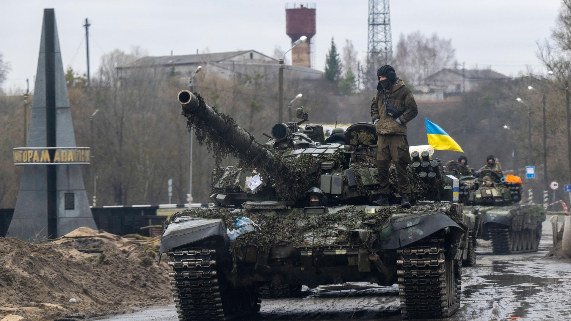 Auf einem Panzer steht ein ukrainischer Soldat, gefolgt von weiteren Panzern. Im Hintergrund ist die ukrainische blau-gelbe Flagge zu sehen.